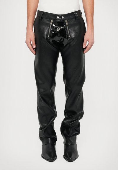 Άνδρας που φοράει μαύρο δερμάτινο παντελόνι με δύο κατακόρυφες μπροστινές φερμουάρ και γυαλιστερή μαύρη λάμψη από δέρμα, συνδυασμένο με μαύρες μυτερές μπότες.
