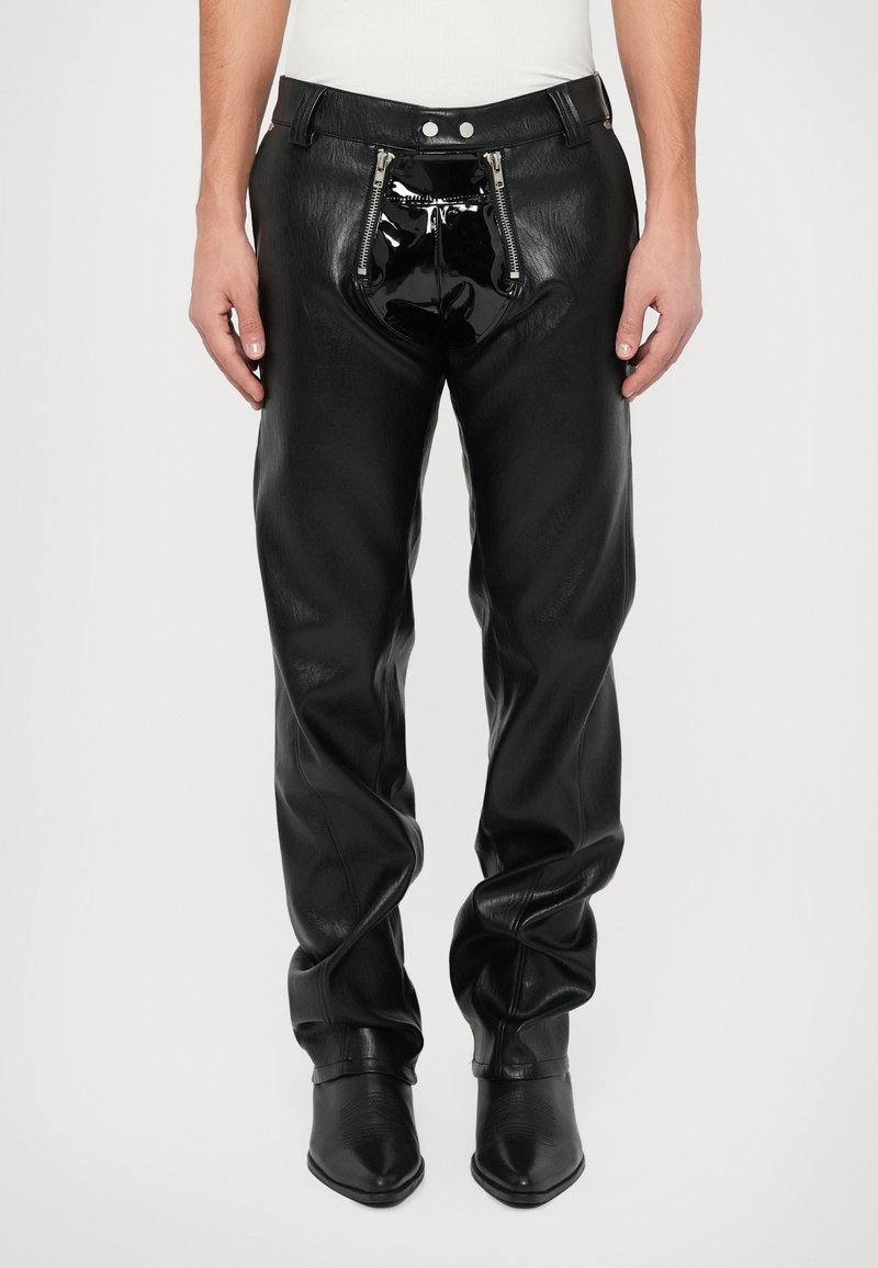 Man in zwarte leren broek met twee verticale ritsen aan de voorkant en glanzend zwart lakleer detail, gecombineerd met zwarte puntige laarzen.