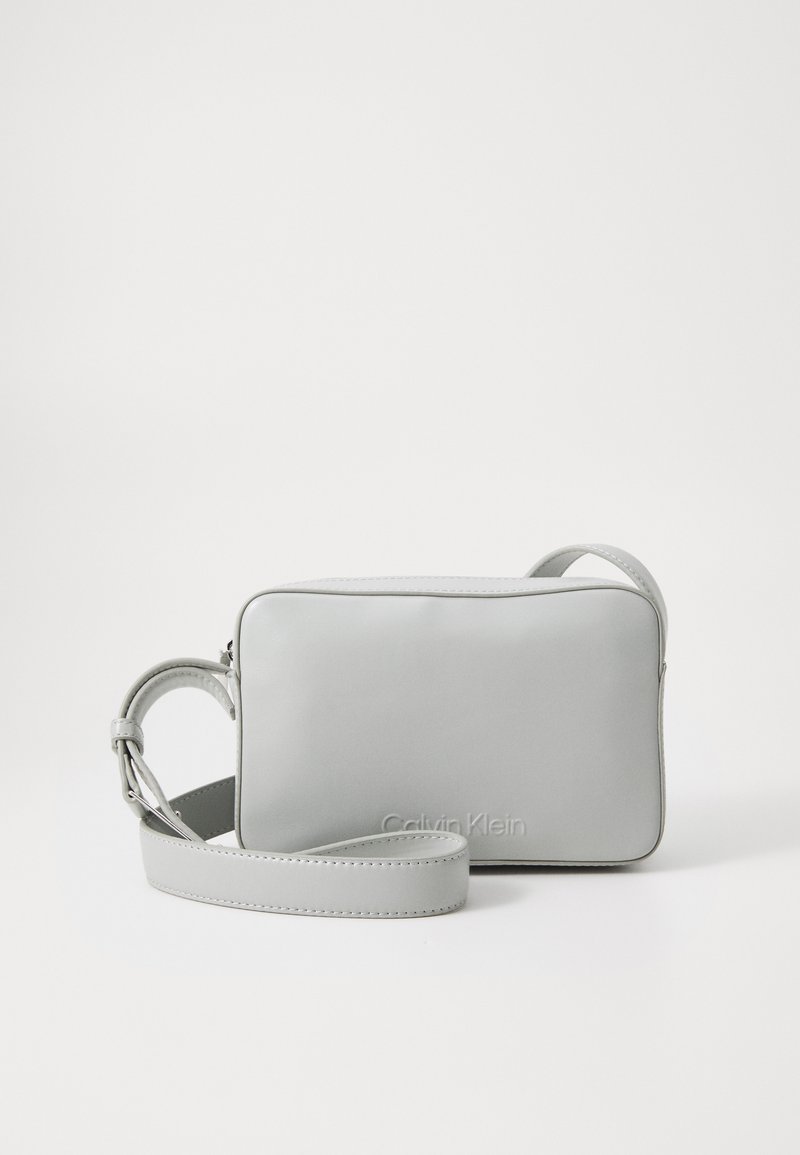 Sivá kožená kabelka cez telo s obdĺžnikovým tvarom, so zipsom a nastaviteľným popruhom. Na prednej strane je vyrazené logo "Calvin Klein".
