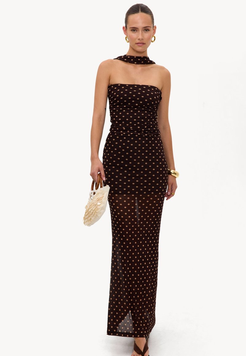 Femme portant une robe bustier noire et marron à pois, tenant un petit sac à main beige avec des poignées en bois sur fond blanc.
