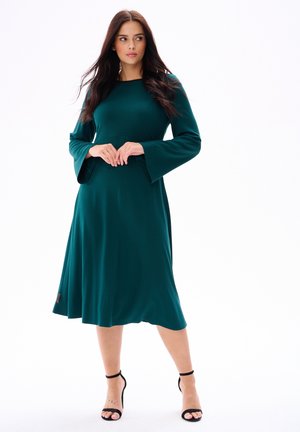 Teal jurk van middellange lengte met een aansluitend lijfje en uitlopende klokmouwen. Soepele textuur, minimalistisch ontwerp en een klein merkteken op de zoom.