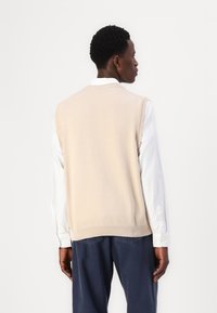 Μπεζ πλεκτό γιλέκο με στρογγυλή λαιμόκοψη, με ριμπ στα άκρα. Φοριέται πάνω από ένα άσπρο μπλουζάκι με μακρύ μανίκι, η πίσω όψη επιδεικνύει τον συνεχόμενο σχεδιασμό.