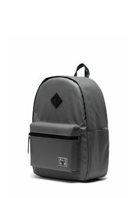 Sac à dos gris en matériau lisse avec une forme arrondie. Comprend une poche avant zippée, des accents noirs et un patch en forme de diamant.