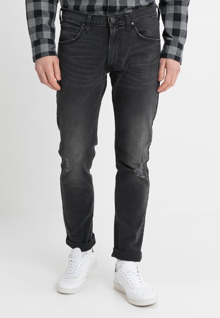 Homme portant un jean slim noir légèrement usé aux genoux, des baskets blanches et une chemise à carreaux noire et grise, debout.