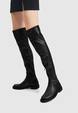 Bottes en cuir noires au-dessus du genou avec fermetures éclair latérales portées avec une jupe noire sur un fond gris clair.