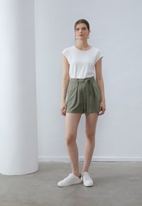 Tricou alb cu mâneci scurte, asortat cu pantaloni šort de culoare verde măsliniu, cu talie înaltă și nod în față. Modelul poartă adidași albi, stând în fața unui fundal deschis.