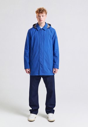 Junger Mann mit lockigem Haar, der einen blauen Kapuzenmantel, dunkle Jeans und weiße Sneakers trägt, steht vor einem einfarbigen hellen Hintergrund.