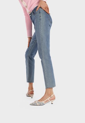 Scarpe slingback a punta con base beige e motivo floreale blu. La modella indossa jeans in denim chiaro con orlo sfrangiato e un pullover rosa a coste.