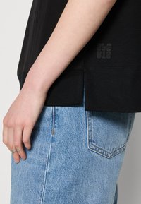 Camiseta de algodón negra con aberturas laterales, con un sutil estampado de logo cerca del dobladillo. Combinada con unos vaqueros de mezclilla azul claro.
