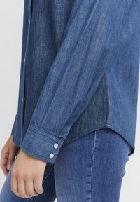 Camicia di denim con polsini abbottonati, orlo leggermente curvo e tessuto testurizzato di un blu medio.