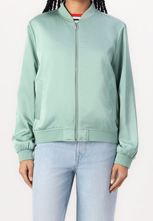 Personne portant une veste bomber vert clair zippée avec poignets et ourlet côtelés, associée à un jean bleu clair.
