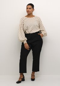 Krämfärgad blus med svarta prickar, elastiska ärmslut; kombinerad med högmidjade svarta byxor och svarta blockklackskor. Enkelt, figursytt design.