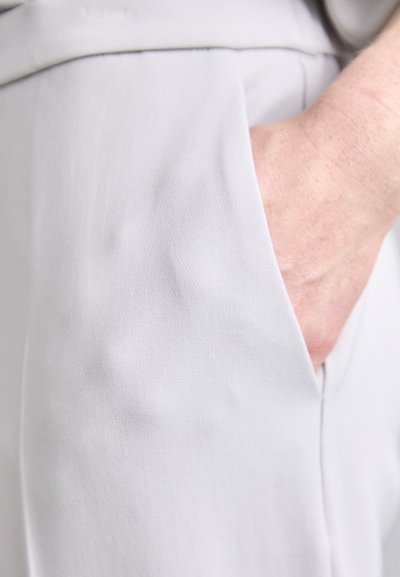 Tessuto grigio chiaro con una superficie liscia, caratterizzato da una tasca laterale e un primo piano di una mano parzialmente visibile al bordo.