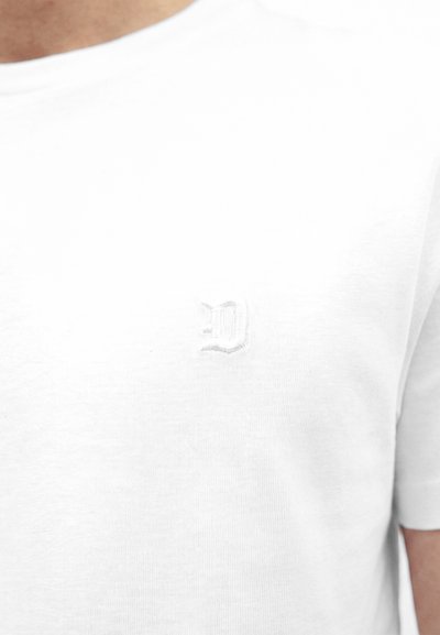 Gros plan d'un t-shirt blanc avec une lettre brodée discrète "D" sur la partie gauche de la poitrine.