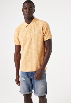 Polo de manga corta en naranja claro con un patrón de hojas, que cuenta con un cuello y un pequeño parche de logo, combinado con pantalones cortos de mezclilla.