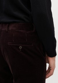 Burgundi színű kordbársony nadrág, texturált felülettel, hátsó zsebbe és gomb részletet tartalmaz. Látható derékpánt és oldalsó varrás.
