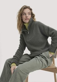 Grauer Reißverschluss-Pullover mit gerippter Textur, kombiniert mit hellgrünen Cordhosen. Der Pullover hat einen hohen Kragen und eng anliegende Bündchen. Sitzt auf einem Holzstuhl.