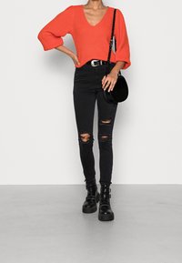 Maglione a maglia oversize color corallo con scollatura a V, abbinato a jeans skinny neri strappati e stivali neri con tacco grosso. Borsa a tracolla nera.