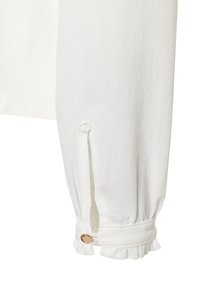 Biała bluzka z długim rękawem, wyróżniająca się teksturowanym materiałem, falbankami przy mankietach oraz guzikiem w okolicy nadgarstka. Dyskretne rozcięcie na rękawie.