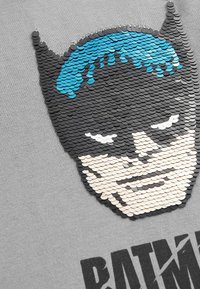 Grauanger T-Shirt aus Stoff mit einem strukturierten, paillettenverzierten Batman-Gesichtsdesign in Schwarz, Blau und Creme, darunter der aufgedruckte Text "BATMAN".