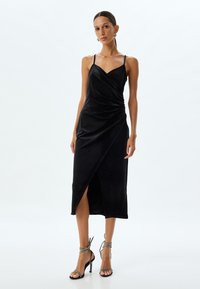 Robe midi en satin noir avec un design drapé, des bretelles fines et une fente sur le côté. Associée à des sandales à talons hauts argentées à lanières.