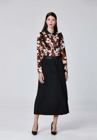 Blouse floral avec un fond brun et un col volanté, associée à une longue jupe plissée noire. Des talons hauts noirs complètent le look.