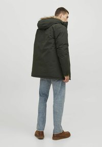 Dark green parka jacket with a fur-lined hood, front zipper, and snap closure; paired with light blue jeans and brown shoes.
