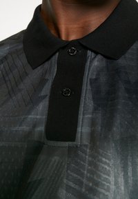 Černé polo tričko s texturovaným vzorem; má dva černé knoflíky a černý límec; vyzdvihuje kombinaci matných a lesklých povrchů.