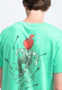 Muntgroene t-shirt met een grafische print van een zittende figuur met een rode appel als hoofd, omringd door pijlen en gebogen lijnen.
