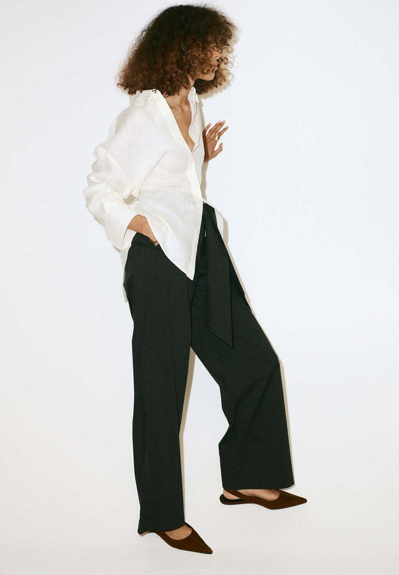 Femme aux cheveux bouclés portant une chemise blanche ample, un pantalon large noir et des chaussures slingback marron à bout pointu, debout de profil.