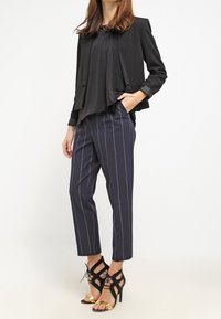 Blazer fluido nero sopra un top nero, abbinato a pantaloni a righe blu navy e sandali neri con tacco alto e accenti dorati.