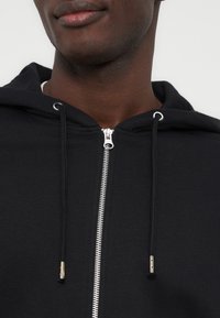 Άνδρας που φοράει μαύρο φούτερ με φερμουάρ και κορδόνια, με εμφανή τον λαιμό και το κάτω μέρος του προσώπου του, σε απλό φόντο.