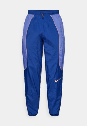 Blauwe nylon trainingsbroek met elastische tailleband, trekkoord en oranje piping. Kenmerkt het Nike-logo aan de linkerzijde. Glad materiaal, tapered fit.
