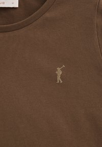 Brun ribbad tyg-topp med en liten guldfärgad nitar-golfare grafik på vänster bröstområde. Normal passform med rund halsringning.