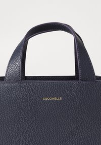 Kožená taška přes rameno v tmavě námořnické barvě s texturovaným povrchem, vybavená dvěma krátkými rukojeťmi a zlatým embosovaným logem na přední straně.