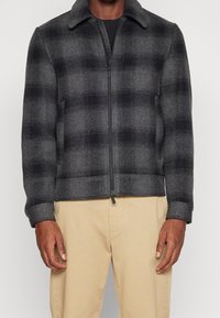 Man i en zip-up svart och grå rutad ulljacka med beige byxor, stående mot en enkel ljus bakgrund.