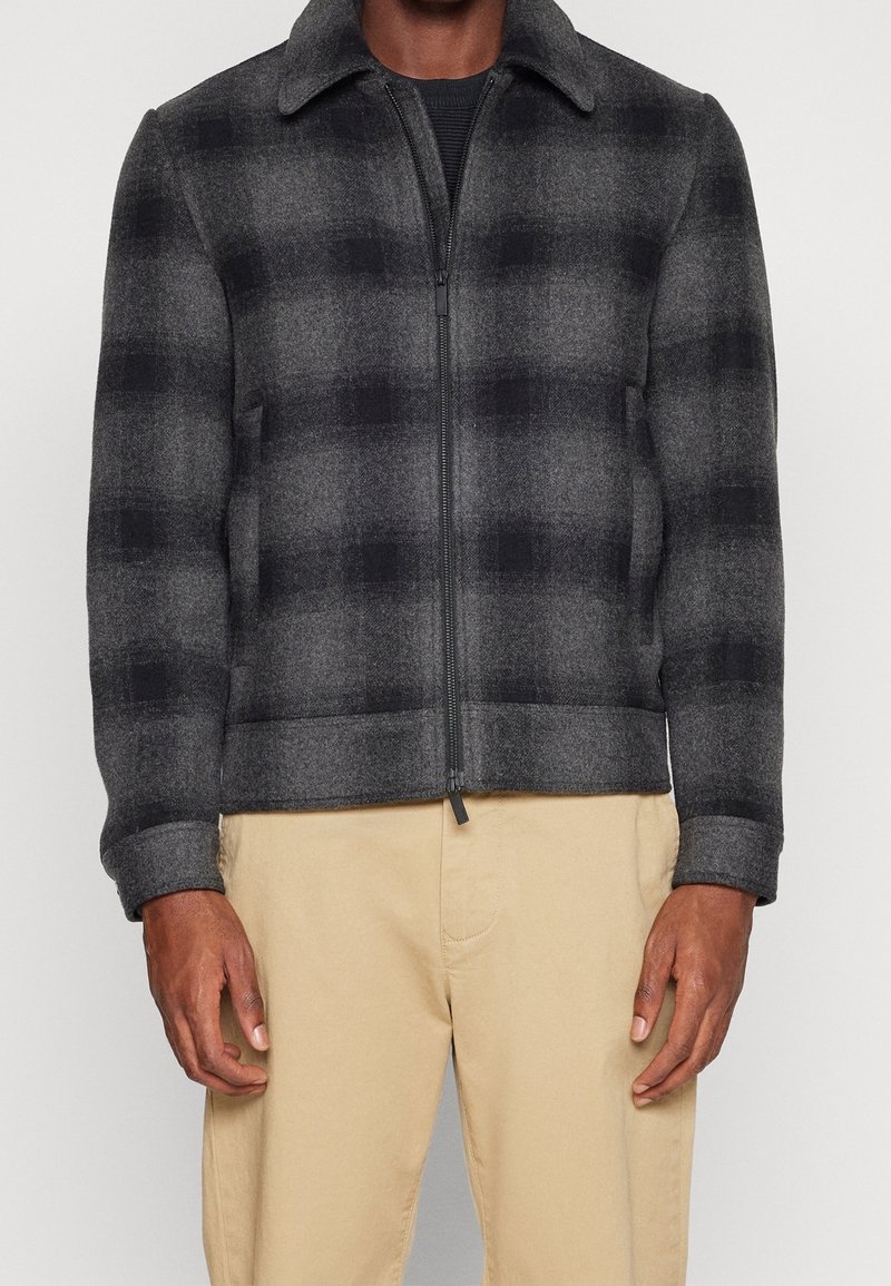 Man i en zip-up svart och grå rutad ulljacka med beige byxor, stående mot en enkel ljus bakgrund.