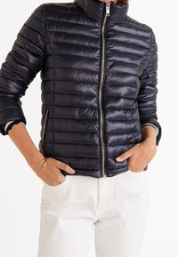 Doudoune noire avec fermeture éclair sur le devant, présentant un design matelassé horizontal et un col montant. Portée par-dessus une chemise et un pantalon de couleur claire.