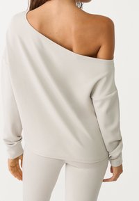 Sweatshirt léger gris clair à épaules dénudées, à manches longues, coupe décontractée, texture douce et détails de couture visibles dans le dos.