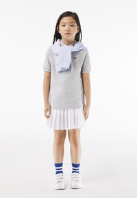 Polo grigio a maniche corte, gonna bianca plissettata, maglione azzurro chiaro annodato attorno al collo, sneakers bianche e calze a righe blu.