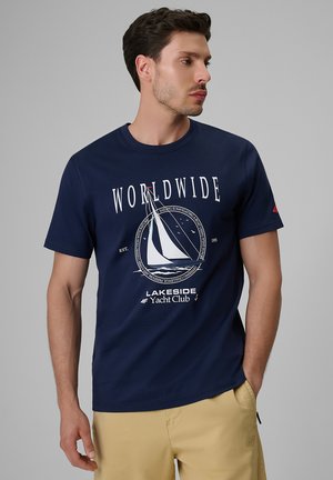 Mężczyzna w granatowym t-shircie z białą grafiką żaglówki i napisem "WORLDWIDE Lakeside Yacht Club", w połączeniu z beżowymi spodniami, stoi na szarym tle.
