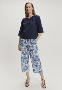 Marinebluse aus Leinen mit elastischen Bündchen, kombiniert mit weißen, taillierten Hosen mit blauen Blumenmustern. Schwarze Riemchensandalen runden den Look ab.