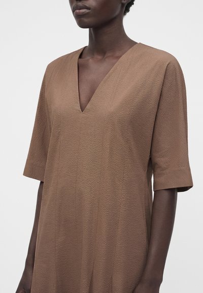 Γυναίκα με καφέ φόρεμα σε σχήμα V και μανίκια μέχρι τον αγκώνα, εμφανίζεται από τους ώμους μέχρι τους γοφούς σε ένα απλό λευκό φόντο.