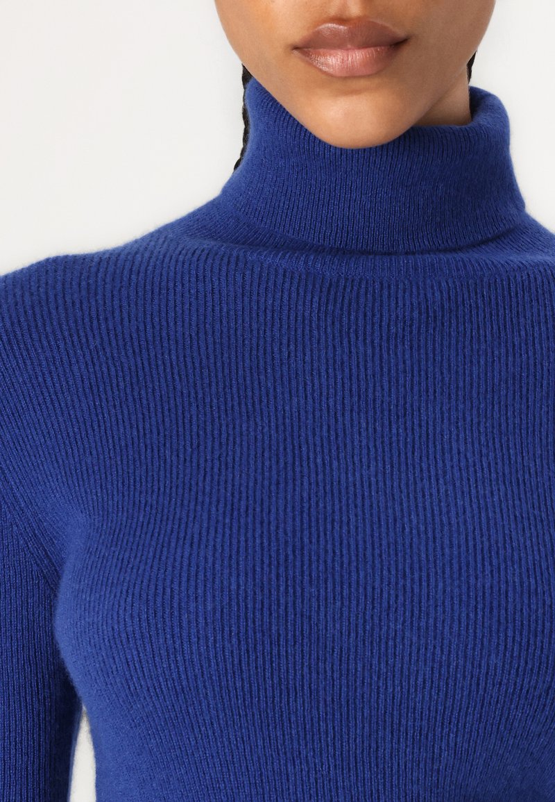 Suéter azul de cuello alto y acanalado con un diseño ajustado. La tela parece suave y texturada, con un cuello alto y mangas largas.