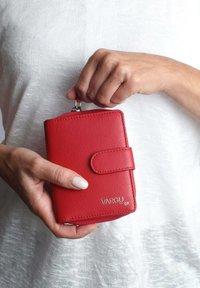 Rote Lederbörse mit glatter Oberfläche, rechteckiger Form und einem magnetischen Druckknopfverschluss. Mit einem silbernen Logo auf der Vorderseite.