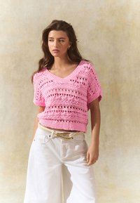 Pink knitted sweater with a V-neck, featuring openwork patterning and short sleeves, paired with wide white trousers and a light belt.