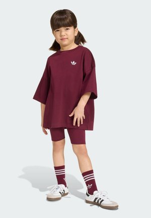Nuori lapsi, jolla on ruskeat lettipidot, pukeutuneena ylisuurikokoiseen marooninväriseen Adidas-paitaan, sopiviin shortseihin, marooninvärisiin sukkiin ja valkoisiin lenkkareihin, seisoo vaalealla taustalla.