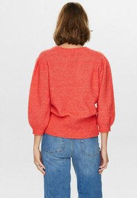 Maglione texturizzato di colore corallo con maniche a sbuffo e orlo a costine. Abbinato a jeans in denim blu. La vista posteriore mostra la vestibilità e il design delle maniche.