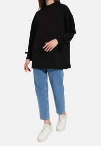 Felpa nera oversize con spalle scese, colletto a coste e dettagli con lacci ai polsini, abbinata a jeans blu chiaro e sneaker bianche.