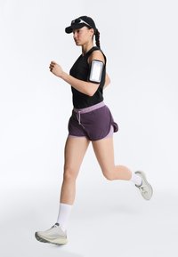 Kobieta biegnąca w czapce z daszkiem, bezrękawniku, fioletowych szortach, białych butach i opasce na ramię na telefon na jednolitym białym tle.